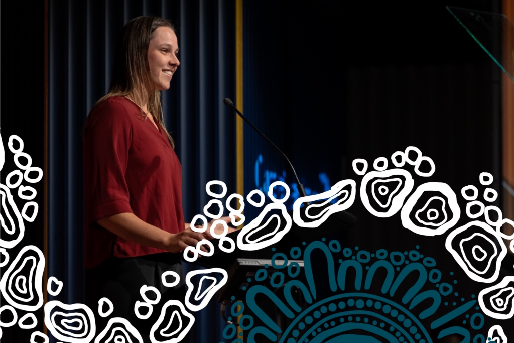 A presenter speaking from a lectern on stage at a CareerTrackers event, framed by First Nations dot-and-track artwork.